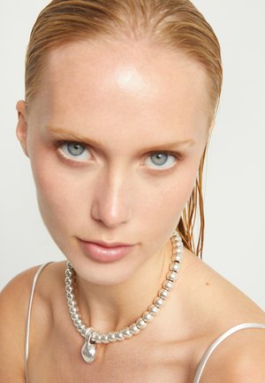 Gros plan sur une femme aux cheveux blonds mouillés portant un collier argenté orné de perles avec un pendentif et un top à fines bretelles blanches.