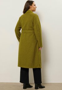 Mujer de pie de lado vistiendo un abrigo largo de tela texturizada en color verde oliva con cinturón, pantalones negros y botines de tacón negro contra una pared clara.