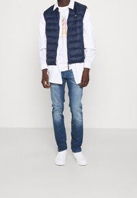 Hombre con chaleco acolchado azul oscuro sobre camisa blanca y camiseta con gráfico, jeans azules y zapatillas blancas, de pie frente a un fondo liso.