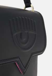 Bolso sintético negro con diseño de ojo en relieve, tapa angular y detalle de acento rosa a lo largo del borde. Adornos en oro.