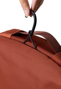 Borsa di tessuto marrone con trama, con un gancio in metallo nero attaccato a una corda; mano posizionata sopra la maniglia superiore della borsa.