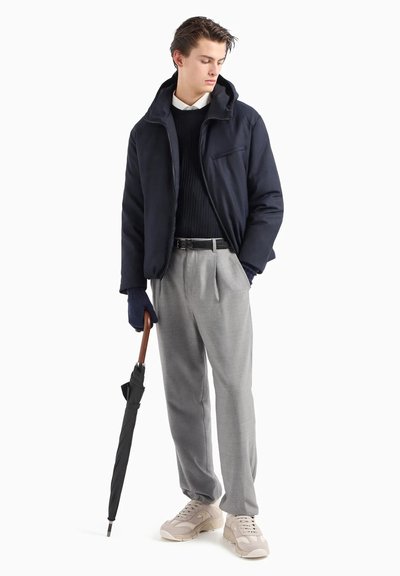 Giacca navy con cappuccio, maglione a coste navy, pantaloni grigi plissettati, sneakers beige e ombrello nero. Tessuto纹otto e design a strati.