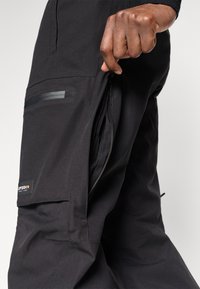 Černé cargo kalhoty s boční kapsou na zip, elegantní látka s jemnou strukturou a viditelnou nášivkou loga na přední straně.