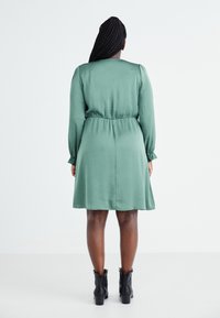 Vihreä, teksturoitu mekko, jossa on vyötäröä korostava leikkaus ja pitkät hihat, ulottuen polven ylle. Yhdistetty mustiin nilkkabootseihin.