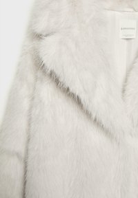 Cappotto in pelliccia ecologica bianca con una texture morbida e soffice, caratterizzato da un colletto a revers e fodera interna. Etichetta visibile nella zona del collo.