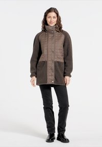 Braune gesteppte Jacke mit strukturierten Fleece-Ärmeln und Taschen. Hoher Kragen und Druckknopfverschluss. Schwarze Hose und Stiefel vervollständigen das Outfit.