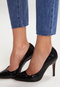 Schwarze, glänzende Lackleder-Pumps mit spitzem Zehenbereich und Stilettosohle. Kombiniert mit blauen, verkürzten Jeans mit ausgefranstem Saum.