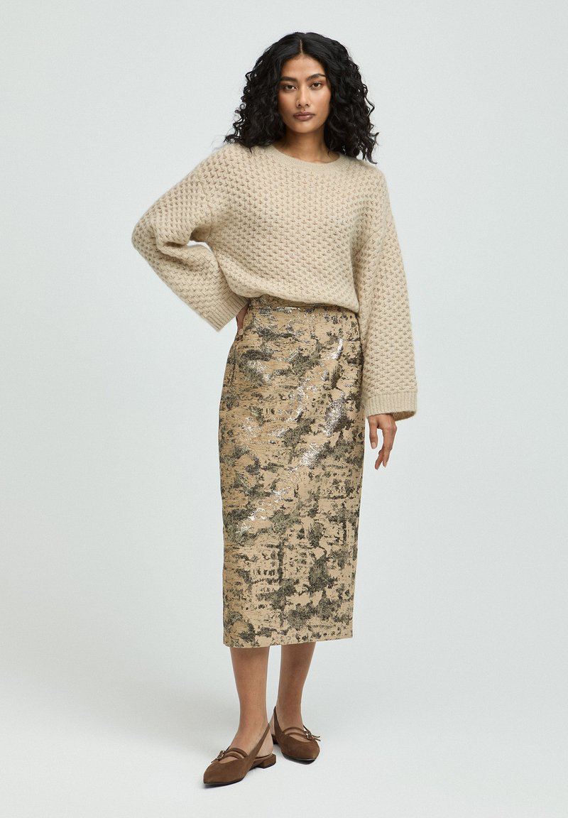 Beige gestrickter Pullover mit überschnittenen Schultern, kombiniert mit einem metallic gemusterten Midi-Rock in gold- und grüntönen. Braune flache Schuhe runden das Outfit ab.
