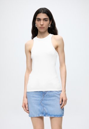 Tânără cu păr lung și închis la culoare poartă un top alb fără mâneci și o fustă din denim bleu deschis, stând în fața unui fundal simplu și deschis la culoare.