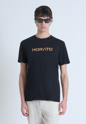 Crna majica kratkih rukava s "MORATO" logotipom u metalik zlatnoj boji, okrugli izrez, meka tekstura, nosi se s tamnim naočalama. Svijetla pozadina.