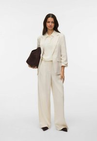 Garnitur w kolorze kremowym z szerokimi spodniami i dopasowaną marynarką, zestawiony z jasną koszulą i ciemną torebką. Gładka tkanina, minimalistyczne detale.