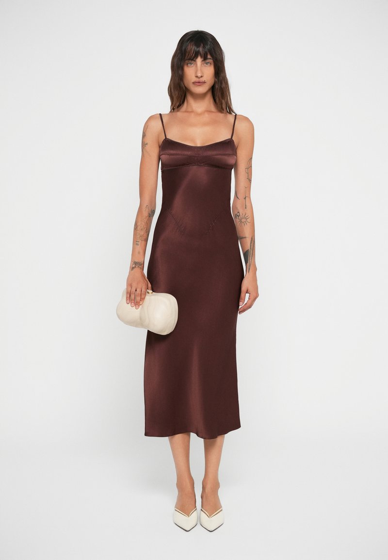 Burgundy satin slip dress with thin straps and a fitted silhouette, paired with a cream clutch and white pointed-toe heels.