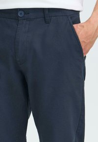 Pantalon bleu marine en tissu lisse, avec une coupe plate, fermeture à bouton et poches latérales. Détails de couture subtils sur l'ensemble.