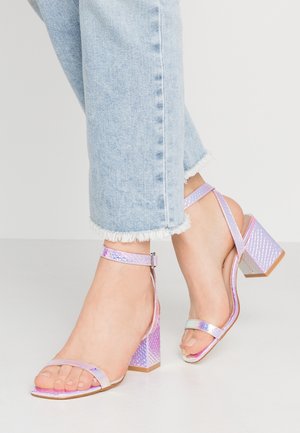 Fødder iført lyse lilla iriserende højhælede sandaler med ankelremme kombineret med lyseblå jeans med frynset kant.