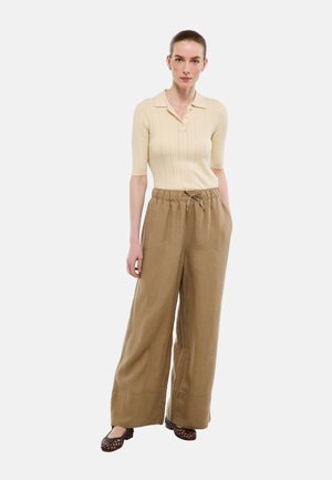 Femme debout portant un polo beige côtelé à manches courtes, un pantalon large marron à cordon de serrage, et des ballerines tissées foncées sur fond blanc.