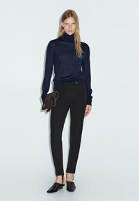 Maglione a collo alto blu navy, pantaloni neri aderenti con bottone, texture liscia e scarpe slip-on nere. La modella tiene una pochette in suede marrone.