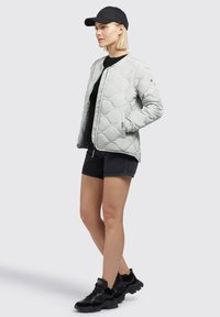 Lichtgrijze gewatteerde bomberjack met elastische manchetten, voorrits en zijzakken. Gecombineerd met zwarte shorts en zwarte sneakers. Zwarte pet op het hoofd.