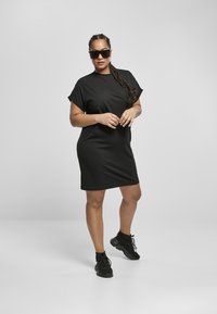 Robe t-shirt noire à manches courtes, coupe décontractée, en tissu lisse. Associée avec des bottines noires et des lunettes de soleil carrées.