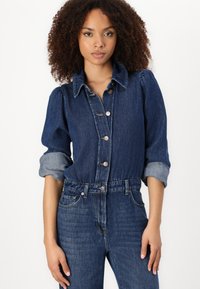 Denim jumpsuit med knappåpning foran, puffede ermer og rullede mansjetter. Stoffet er mørkeblått med en glatt tekstur og subtil falming.