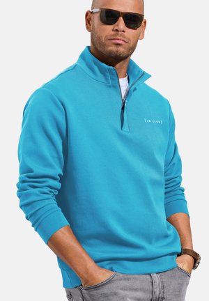 Mann trägt schwarze Sonnenbrille und ein blaues Viertel-Reißverschluss-Sweatshirt mit "JD Club" Logo, Hände in den Taschen und graue Hose.
