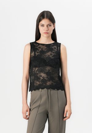 Black lace sleeveless top with floral pattern, scalloped hem, semi-transparent design, paired with olive-green trousers.