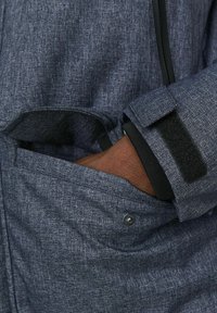 Tejido de chaqueta gris texturizado, que presenta un bolsillo lateral con solapa, con una mano parcialmente visible dentro. Cierre de cinta de velcro en el puño.