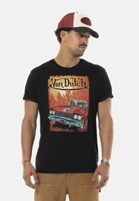 T-shirt noire ornée d'un graphique coloré d'une voiture vintage avec des flammes et le texte "Von Dutch". Le mannequin porte une casquette de baseball rouge et blanche.