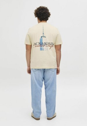 Beige T-Shirt mit einem Grafikdesign eines Wolkenkratzers und Text auf dem Rücken. Getragen mit hellblauen Jeans und beigen Sneakers.