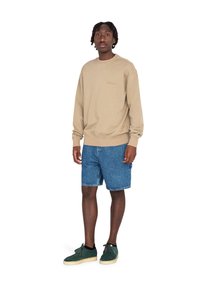 Sudadera beige con cuello redondo, pantalones cortos de mezclilla azul y zapatos de ante verdes. El modelo adopta una postura neutra frente a un fondo blanco.