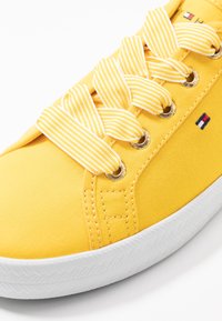 Żółty materiałowy tenisówka z białą gumową podeszwą, wyposażona w złote przelotki i żółte sznurowadła w paski, z logo marki po boku. Gładka tekstura.