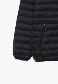 Svart puffjacka med quiltad textur, som har en åtsittande mudd och en dragkedjeficka på sidan. Mjuk, lätt material.