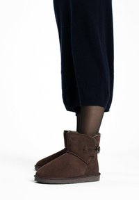 Botas de tobillo de ante marrón con un forro suave, puntera redonda y detalle de botón, combinadas con pantalones de punto oscuros.