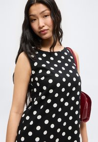 Γυναίκα με μακριά μαύρα μαλλιά φοράει μαύρο αμάνικο φόρεμα με λευκές βούλες και κρατάει μια κόκκινη τσάντα ώμου.