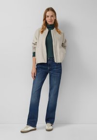 Beige bomberjack, groene coltrui, blauwe spijkerbroek, witte sneakers, met ribgebreide manchetten en een gladde textuur. Neutrale achtergrond.