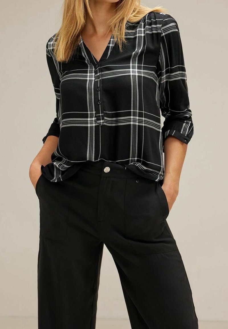 Camicia a quadri bianco e nero con scollo a V, maniche lunghe arrotolate ai polsini, indossata con pantaloni neri. Tessuto di un aspetto liscio.