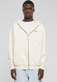 Cream zip-up hoodie in soft fabric, featuring a kangaroo pocket, ribbed cuffs, and a signature logo on the left chest.