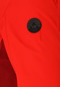 Rote isolierte Skihosen mit glatter Textur, ausgestattet mit einem runden schwarzen Logopatch mit Bergdesign an der oberen Oberschenkelpartie.