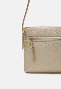 Metalická zlatá crossbody taška vyrobená z texturované umělé kůže. Obsahuje horní zipové zavírání a kovové detaily v zlaté barvě s třásněmi na zipu.