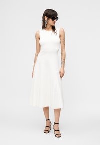 Γυναίκα με τατουάζ που φοράει λευκό πλισέ μίντι φόρεμα χωρίς μανίκια, μαύρα ψηλοτάκουνα σανδάλια, μεγάλα μαύρα γυαλιά ηλίου και μακριά σκουλαρίκια.