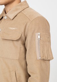 Beżowa kurtka z korduroju z kołnierzykiem, kieszenią na piersi i zamkniętą kieszenią na rękawie. Charakteryzuje się pionowym prążkowanym wzorem oraz srebrnym osprzętem.