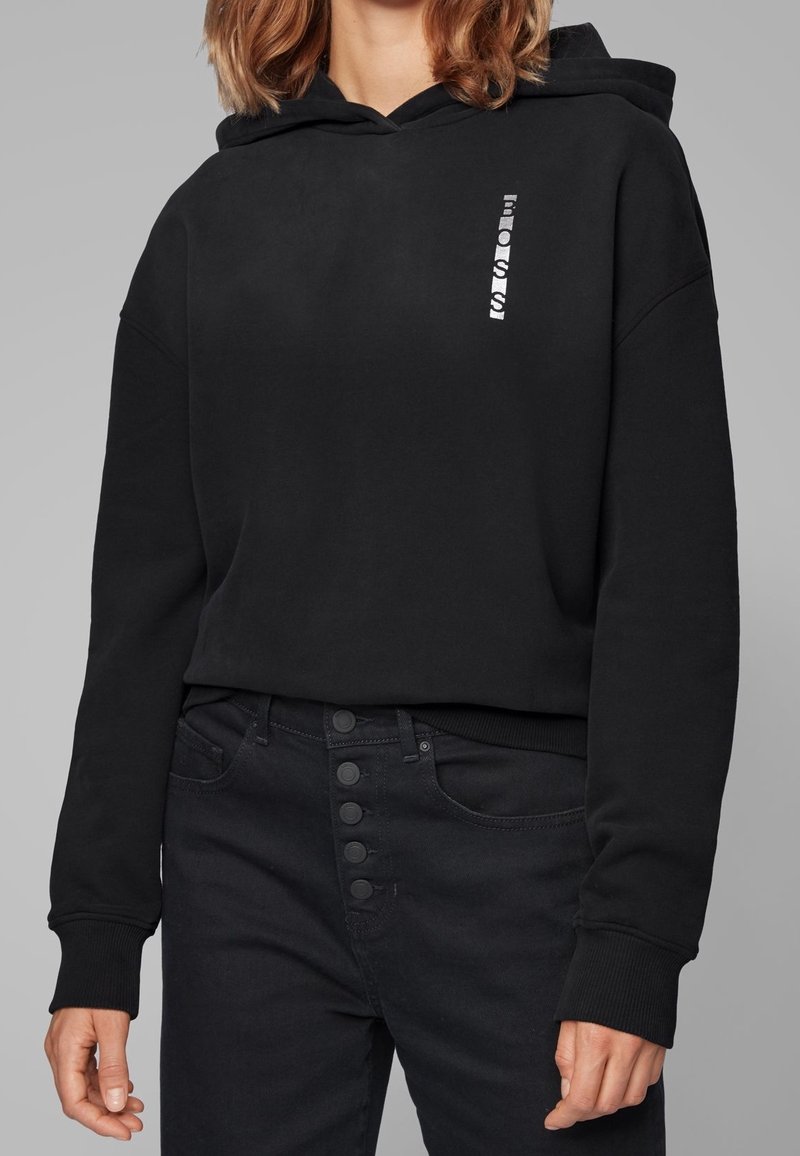 Svart hoodie med dragsko på huvan och en silverfärgad logotyp tryckt vertikalt fram. Matcher med svarta högmidjade jeans med knappstängning.