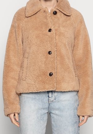 Giacca in pile teddy beige con ampio collo e quattro grandi bottoni scuri, indossata sopra jeans azzurri a vita alta.