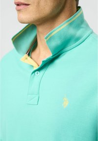Vaaleansininen polo-paita, jossa on keltainen kaulus, kahden napin neppari ja brodeerattu logo. Sujuva rakenne, lyhyet hihat, urheilullinen muotoilu.