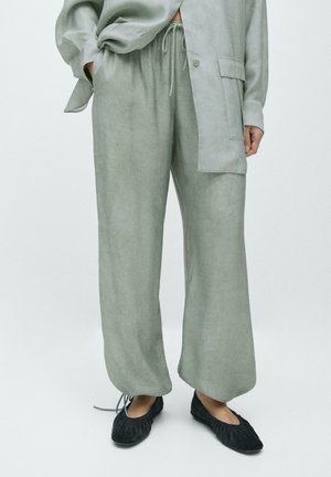 Luźne jasnoszare spodnie z wiązanym pasem, noszone z pasującą koszulą i czarnymi balerinami z marszczonym wykończeniem.