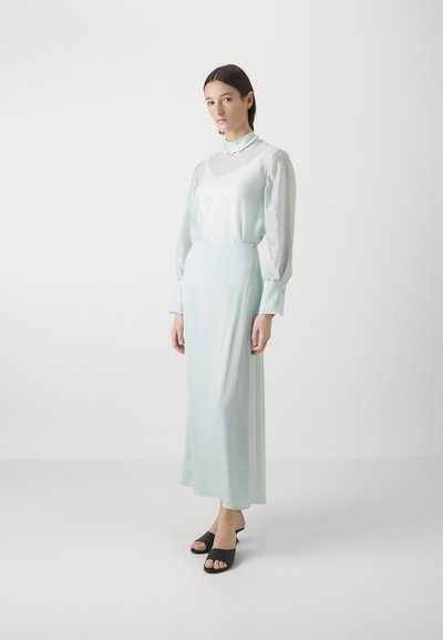 Lungo vestito menta chiaro con maniche lunghe trasparenti, scollatura alta e vita aderente. Abbinato a mule nere open-toe. Design minimalista.