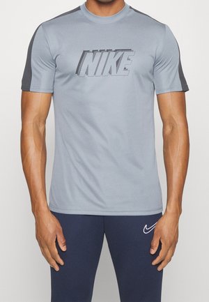 Mann trägt ein hellgraues Nike-T-Shirt mit schwarzen Akzenten und dunkelblauen Nike-Leggings, steht mit entspannten Armen seitlich am Körper.