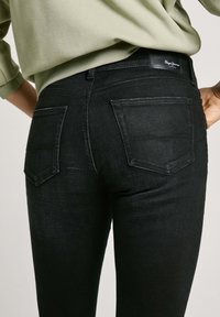 Juoda džinsai, turintys dvi galines kišenes, elegantišką dizainą ir lygų paviršių. Dėvimi su švelnia žalia palaidine, matoma juostos logotipas.