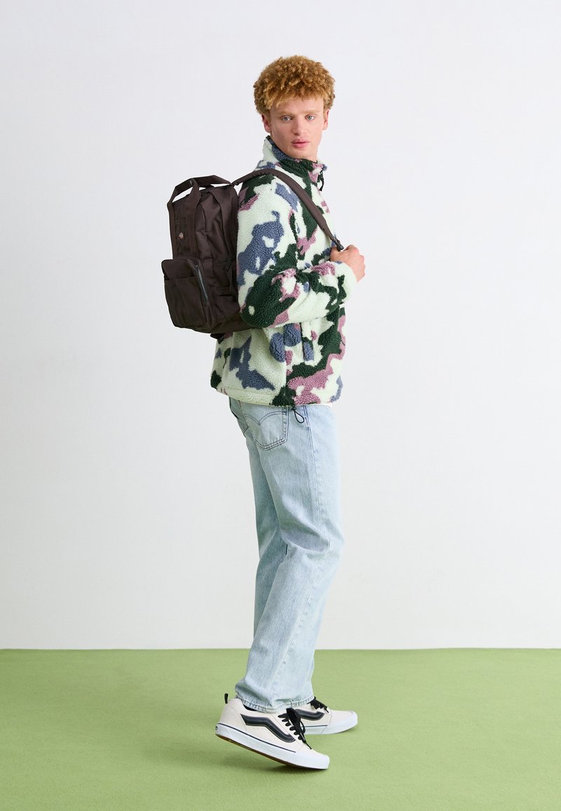 Chaqueta de forro polar con patrón de camuflaje en verde, azul y rosa, combinada con unos jeans de color azul claro y unas zapatillas negras y blancas, llevando una mochila marrón.