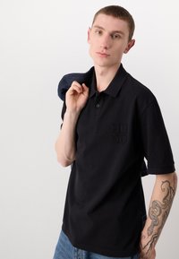 Sort polo-shirt med korte ærmer, der har et præget "HUGO" logo på brystet. Modellen holder en denimjakke over den ene skulder.