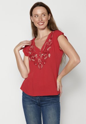 Haut rouge à manches courtes avec un décolleté en V et des détails de broderie florale. Tissu doux avec une coupe décontractée et un ourlet légèrement évasé.